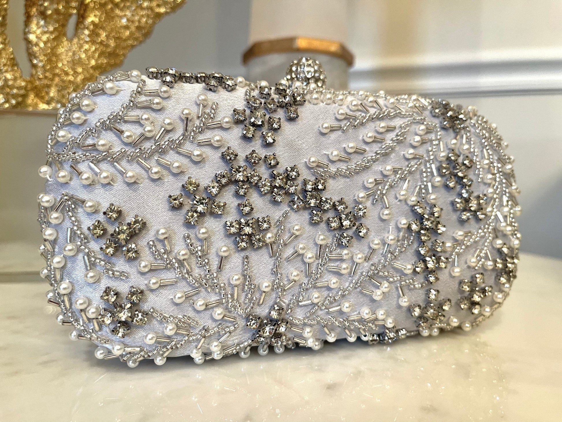 Front view of silver crystal leaves and pearl beads embroidered clutch with gold and white details in the background