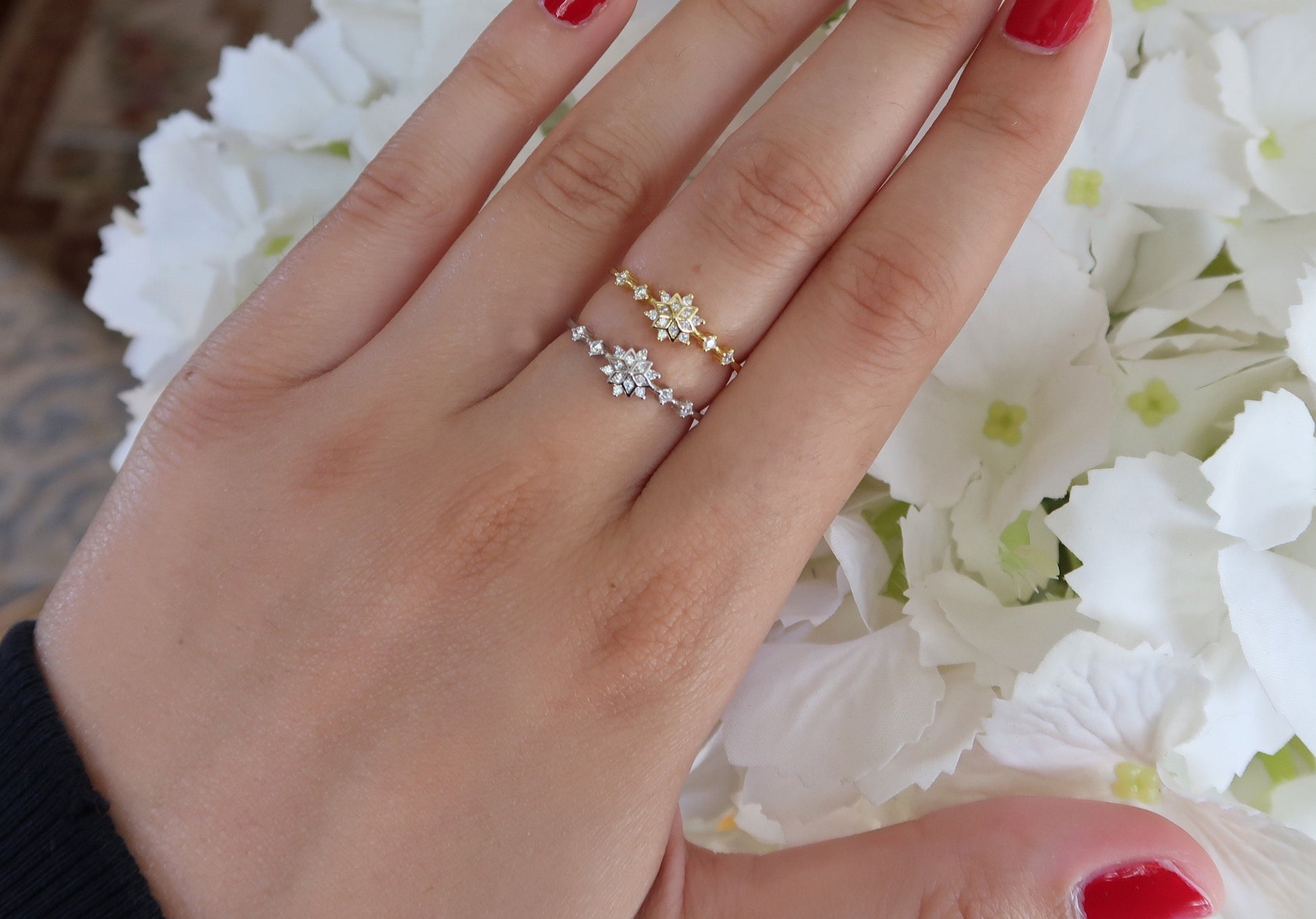 Silver and gold snowflake rings modeled on a hand against a white floral background, demonstrating their delicate and elegant design.